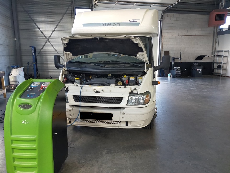 Witte camper met open motorkap, aangesloten op een groen airco-servicestation in de werkplaats